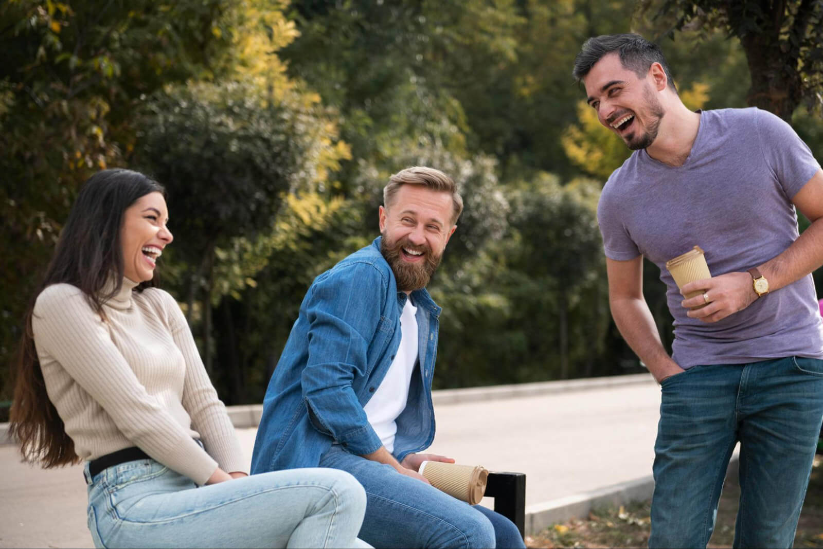 Three young adults sit outdoors in a park, laughing and talking together while holding coffee cups.