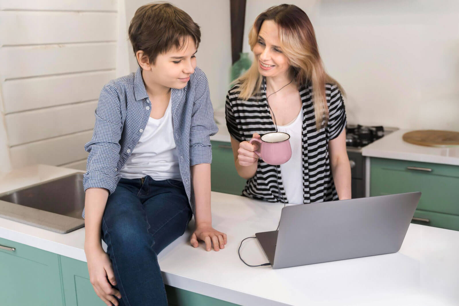 A mother holding a mug smiles at her son as they sit near an open laptop in the kitchen.