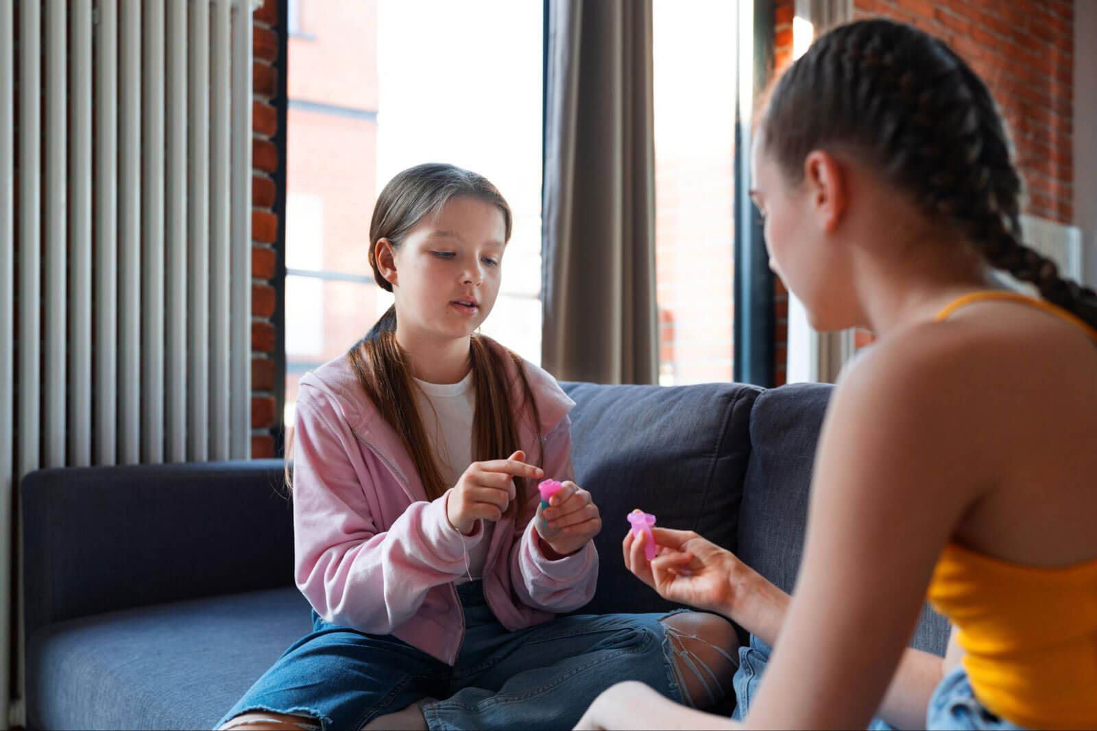 Two young girls sit on a couch talking while holding small toys.