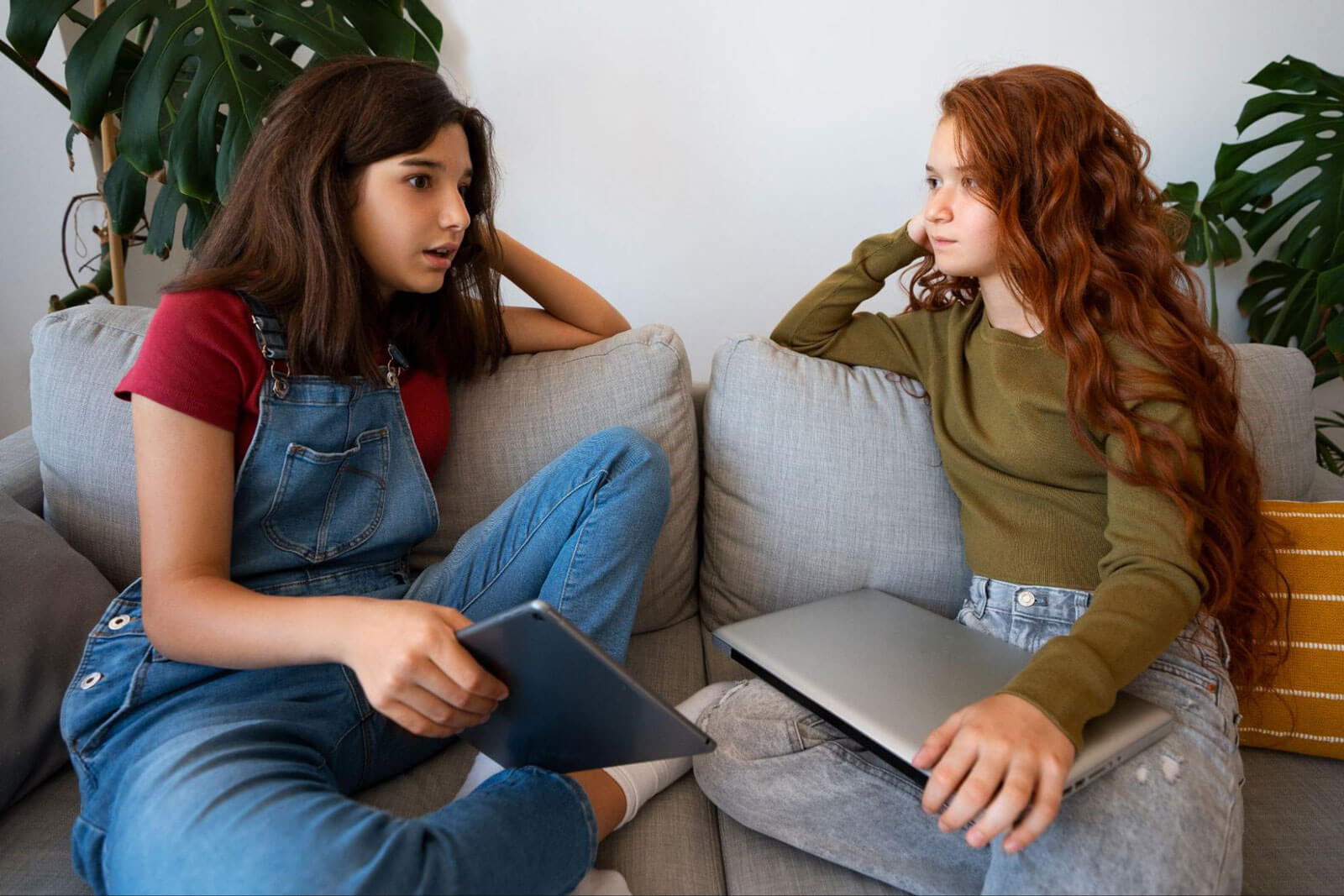 Two teen girls sit on a couch, one talking with a concerned expression while holding a tablet and the other listening with a laptop on her lap.