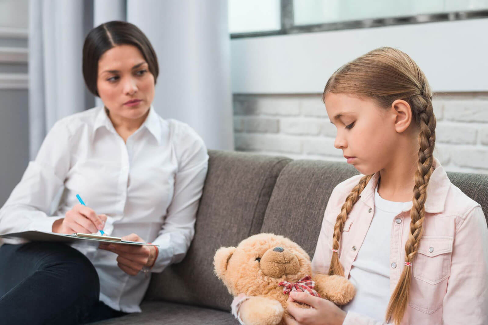 A young girl holding a teddy bear looks down while a counselor takes notes on a clipboard beside her.