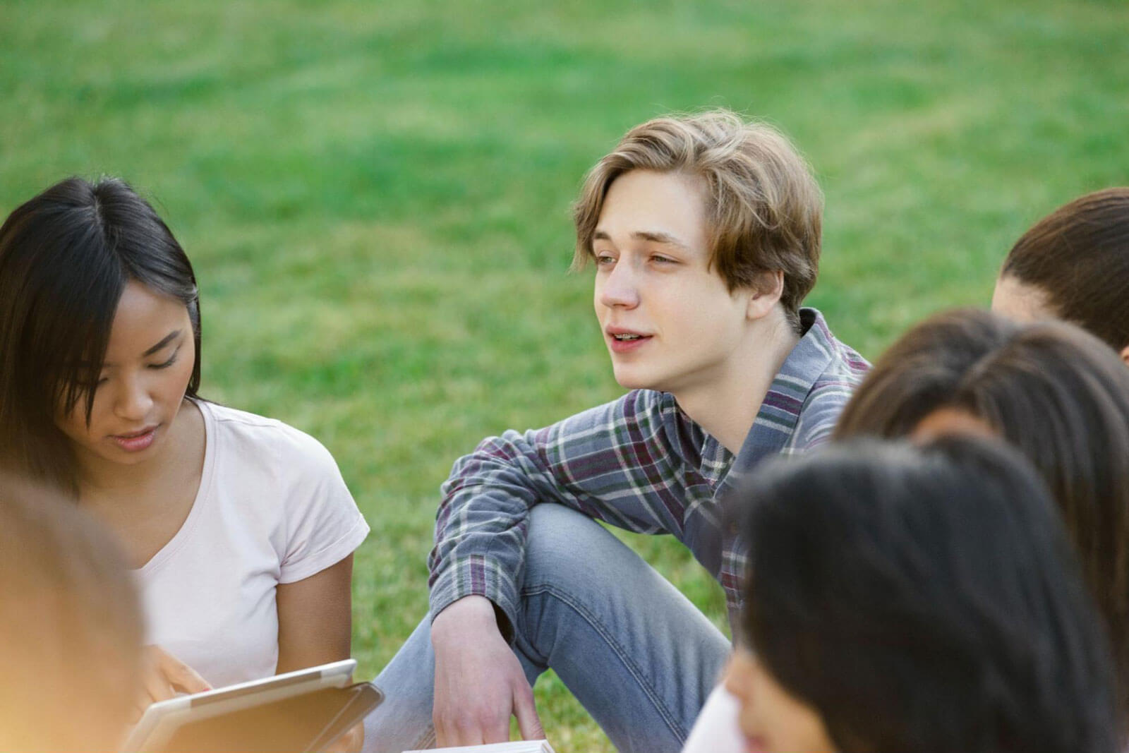 Teenage boy talking to a group of friends sitting together on grass outdoors.
