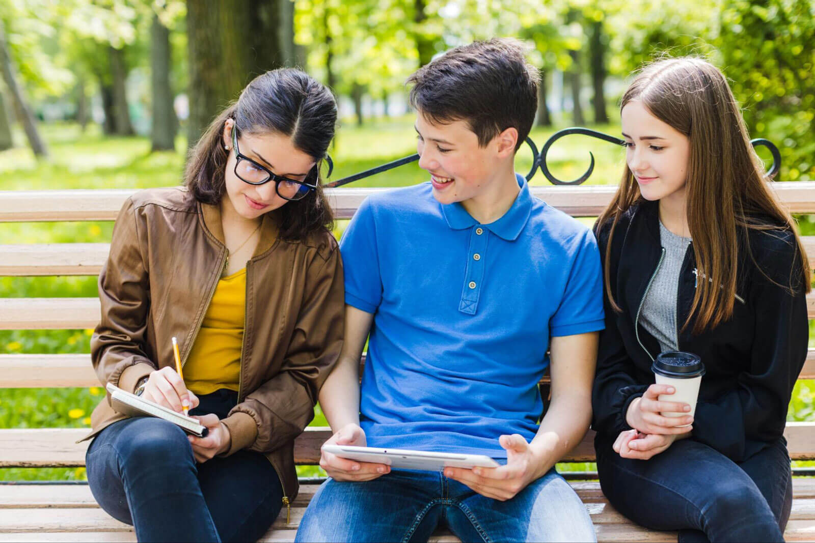 Three teens sitting on a park bench sharing a tablet and coffee together.