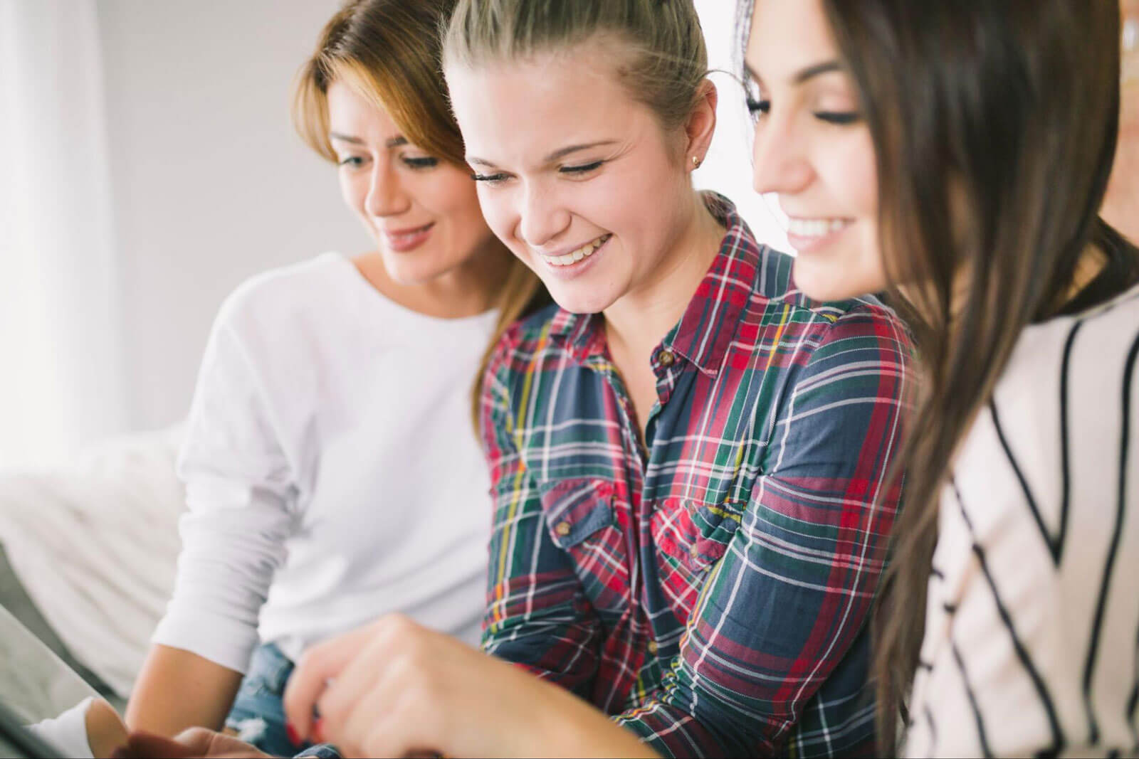 Three smiling women look together at something off-screen while sitting close together.